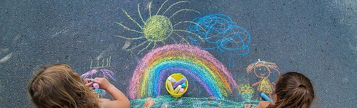 Ein mit Kreide gemalter Regenbogen auf dem Boden, davor zwei Kinderköpfe von hinten